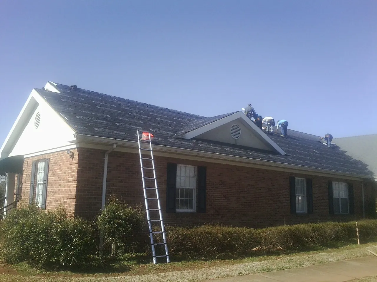 Crew tearing shingles off a brick ranch roof with a ladder set — Emergency Roof Repair work by our crew in Mauston