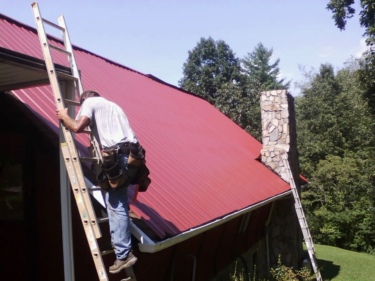 Roofer on a ladder beside a red metal roof and stone chimney — Soffit & Fascia Repair by Roofing Brothers in Mauston